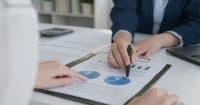 Two people's hands are visible over a desk, analyzing a document featuring financial charts. One hand, wearing a ring, points a finger at a blue and white pie chart with segments labeled "25%" and "66%". The other hand holds a black pen, pointing to a similar blue and white pie chart on the same document. A calculator and a laptop are partially visible in the blurred background.