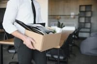 A person in a white button-up shirt, black tie, and dark gray pants is shown from the chest down, holding a light brown cardboard box filled with office items. Contents include black binders, papers, and a small white pot containing a dark green spiky plant. The background shows a blurred modern office interior with wooden desks and a dark grid-patterned shelf.