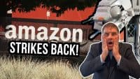 A composite image featuring the 'amazon' logo on a dark fence against a red building, a man in a dark suit with a surprised expression holding his face, and a large grey AT-AT Walker from Star Wars. The text 'AMAZON STRIKES BACK!' in white letters on a black bar overlays the bottom left. Green and silver grasses are in the foreground.