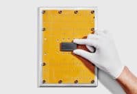 A white-gloved hand delicately places a rectangular black IBM Quantum 'Nighthawk' chip onto a bright yellow circuit board. The board features numerous small holes, larger circular openings, and several silver screws around its edges, all set against a plain white background.