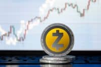 A close-up shot shows a silver Zcash cryptocurrency coin with a yellow core and a black stylized 'Z' logo, standing upright on a stack of two other similar coins. In the blurred background, a digital screen displays a candlestick chart with green and red bars indicating market movements.