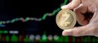 A person's hand holds a golden Zcash cryptocurrency coin. The coin has a large 'Z' symbol and the words 'ZCASH', 'decentralized', 'shielded', 'secured' around its edge. In the background, a blurred digital display shows a financial market chart with green and red lines trending upwards against a dark background, signifying growth.
