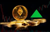 Several golden Ethereum cryptocurrency coins are displayed on a reflective dark surface. A large, prominent Ethereum coin with its logo and 'ethereum' text is in the center-right. In the dark background, a financial candlestick chart shows both green upward trends and red downward movements. A large, bright green triangle, pointing upwards, is positioned above the coins on the right side of the image.