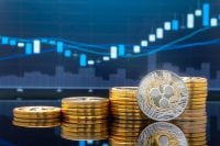 A silver and gold Ripple (XRP) cryptocurrency coin stands upright among several stacks of shiny gold coins, reflecting on a dark, polished surface. In the blurred blue background, a financial chart displays upward-trending candlestick bars and lines, suggesting market growth.