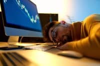 A close-up shot of a man with dark hair and a beard, wearing glasses and a yellow sweater, sleeping with his head resting on a light-colored desk. To his left, a computer monitor displays blue and white financial charts. A keyboard and mouse are visible on the desk.