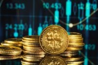 Close-up of several stacks of gold-colored cryptocurrency coins, prominently featuring an XRP coin with its distinct logo and text. The coins are shiny and reflective, sitting on a dark surface. In the blurred background, a digital financial chart with blue glowing lines, bars, and numbers like '10.453' and '13.343' indicates market activity.