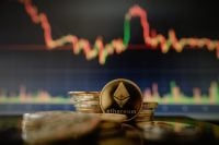 A close-up of a golden Ethereum coin, featuring the Ethereum logo and 'ethereum' text, stacked among other gold-colored coins. In the out-of-focus background, a digital screen displays a financial market chart with fluctuating red and green lines, indicating price changes.