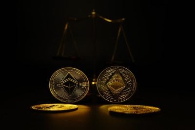 Two golden Ethereum coins stand upright in the center of a dark image, with two more golden coins lying flat in the foreground. In the blurred background, a golden balance scale is visible, all set against a black backdrop with dramatic spotlighting on the central coins.