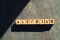 Wooden blocks spelling out "ALTCOINS" are arranged on a textured surface, possibly wood or concrete, with a strong shadow cast across a portion of the image.
