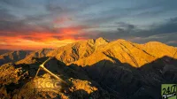 An aerial view of rugged mountains at sunset or sunrise, bathed in warm golden light, with long shadows extending across the terrain. The sky transitions from vibrant orange and red at the horizon to dark blue-grey higher up. In the foreground, on a rocky peak, a golden pickaxe rests, and the letters 'KGC' glow with a golden light embedded in the rock. A '24/7 WALL ST' logo is visible in the bottom right corner.