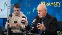 A bald man with a white beard and glasses, wearing a dark blue collared shirt and black blazer, speaks into a silver studio microphone on a boom arm. He holds a black smartphone in his left hand, displaying a bright red sports car parked on an asphalt road. Behind him, a younger man with short brown hair, wearing a light tan hoodie, looks down at his own black smartphone. A clear plexiglass divider separates the two. In the background, large blue screens show financial bar charts in green and red. The yellow 'THE RAMSEY SHOW' logo is prominently displayed on the blue background, and a '24/7 WALL ST' logo is in the bottom right corner.