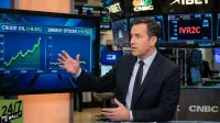 A male news anchor, Brian Sullivan, in a dark suit and tie, stands in a brightly lit financial news studio, gesturing with both hands. Behind him, a large digital screen displays two line graphs: the left shows 'CRUDE OIL (+9.5%)' with a steep upward trend from 10 to 50, and the right shows 'ENERGY STOCKS (+0.1%)' with a relatively flat trend. Various other financial data screens with CNBC and 'XIBET' logos are visible in the background, along with a '24/7 WALL ST' logo in the bottom left corner.