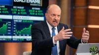 A bald man with a goatee, Jim Cramer, wearing a dark suit and blue tie, speaks animatedly with his hands gesturing upwards. He is in a television studio with large digital screens in the background displaying green stock market charts and financial data. A laptop with a '24/7 WALL ST' logo is visible on a table to his right.