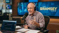 A middle-aged man with a white beard, glasses, and headphones speaks into a black microphone, gesturing with his left hand. He is seated at a dark wooden desk with papers and an open laptop displaying financial charts. In the background, two large blue screens feature financial graphs and 'THE RAMSEY SHOW' logo.