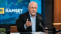 A bald man with glasses and a white beard, wearing a light blue shirt and dark blazer, speaks into a black microphone on a desk. His right hand is raised in a gesture. Behind him is a large blue screen displaying 'THE RAMSEY SHOW' logo and blurred financial charts. A green '24/7 WALL ST.' logo is visible in the bottom left corner.