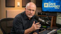 A bald man with glasses and a grey beard, Dave Ramsey, is seated at a desk in a radio studio, speaking into a silver microphone. He is wearing a dark shirt and gesturing with his right hand. Behind him, a television screen displays 'The Dave Ramsey Show' logo in blue and yellow.