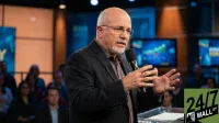A bald man with a gray beard and glasses, wearing a dark suit jacket over a patterned shirt, speaks into a black microphone while gesturing with his hands. He stands behind a podium with papers. The background features blurred studio lights, blue screens displaying financial charts and text, and a seated audience. A '24/7 WALL ST' logo is visible in the bottom right corner.