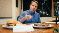 A man in a blue button-down shirt sits at a wooden table in a podcast studio, gesturing with his hand as he speaks into a large professional microphone. In front of him are two white plates: one holds a cheeseburger with lettuce, tomato, and cheese, accompanied by a side of fries; the other contains a grilled steak with a green garnish. Several coins and a white paper document, resembling a check, are scattered on the table.