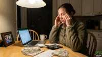 A woman in a military green fleece jacket with a "MILITARY" patch sits at a wooden kitchen table, hands on her temples, eyes closed in distress. On the table are stacks of U.S. dollar bills, a silver laptop showing a financial spreadsheet, a black calculator, white papers, and a coffee mug. A framed photo of two smiling children is to her left. The background shows a dimly lit kitchen with white cabinets.