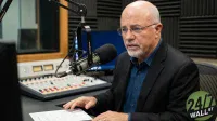 A bald man with glasses and a white beard, wearing a dark suit jacket and blue collared shirt, sits in a radio studio speaking into a large black microphone on a boom arm. In the background, a sound mixing board with numerous colorful sliders and knobs is visible, along with a window looking into another room and black acoustic foam panels on the walls. A '24/7 WALL ST' logo is in the bottom right corner.