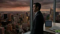 A man in a dark suit stands in profile, looking out a large window at a sprawling cityscape under a cloudy orange and grey sky at dusk. City lights twinkle below, and on a wooden table to the right, a dark tablet stands next to a newspaper featuring the headline 'Oil Spikes'.
