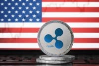 A silver Ripple (XRP) cryptocurrency coin with a blue logo sits on a stack of coins. Behind it, a blurred American flag with red and white stripes and a blue field of white stars is visible.