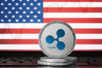 A silver Ripple (XRP) cryptocurrency coin with a blue logo sits on a stack of coins. Behind it, a blurred American flag with red and white stripes and a blue field of white stars is visible.