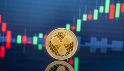 A close-up, golden XRP cryptocurrency coin stands upright on a reflective dark surface. In the blurred background, a digital candlestick chart displays red and green bars against a dark blue grid, indicating market price movements.