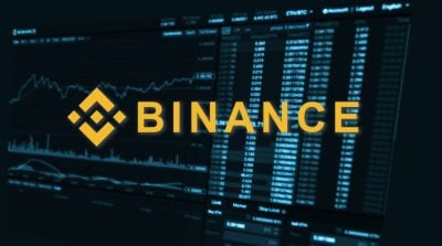 A golden, abstract geometric logo resembling a knot and the word 'BINANCE' in large, bold golden letters are centered on a dark blue digital screen. The background displays blurred cryptocurrency trading data, including price lists, amounts, and a line graph, rendered in light blue and white text, suggesting a busy financial interface.