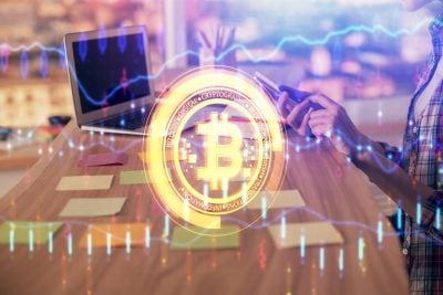 A vibrant, digitally enhanced photo shows a person in a plaid shirt using a tablet at a wooden desk, next to a laptop. A glowing, golden Bitcoin logo with surrounding text like 'DIGITAL CRYPTOGRAPHY' is superimposed over the scene. Blue and purple digital financial graphs and bar charts are overlaid across the entire image, illustrating market data and technology.