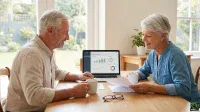 A smiling older man and woman sit opposite each other at a light wooden table, looking engaged as they review financial documents and a laptop displaying a 'Portfolio Dashboard' with various charts and graphs. Both hold white mugs, and a pair of reading glasses rests on the table between them. Large windows in the background reveal a green garden.