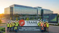 A group of Trane Technologies employees in hard hats and safety vests stands in front of their glass-paneled headquarters building, which features solar panels on the roof. A large company sign and a digital display showing 'Job Growth' charts are visible. The employees hold a banner that reads 'TRANE TECHNOLOGIES - 45,000+ EMPLOYEES & GROWING', set against a warm sunset backdrop.
