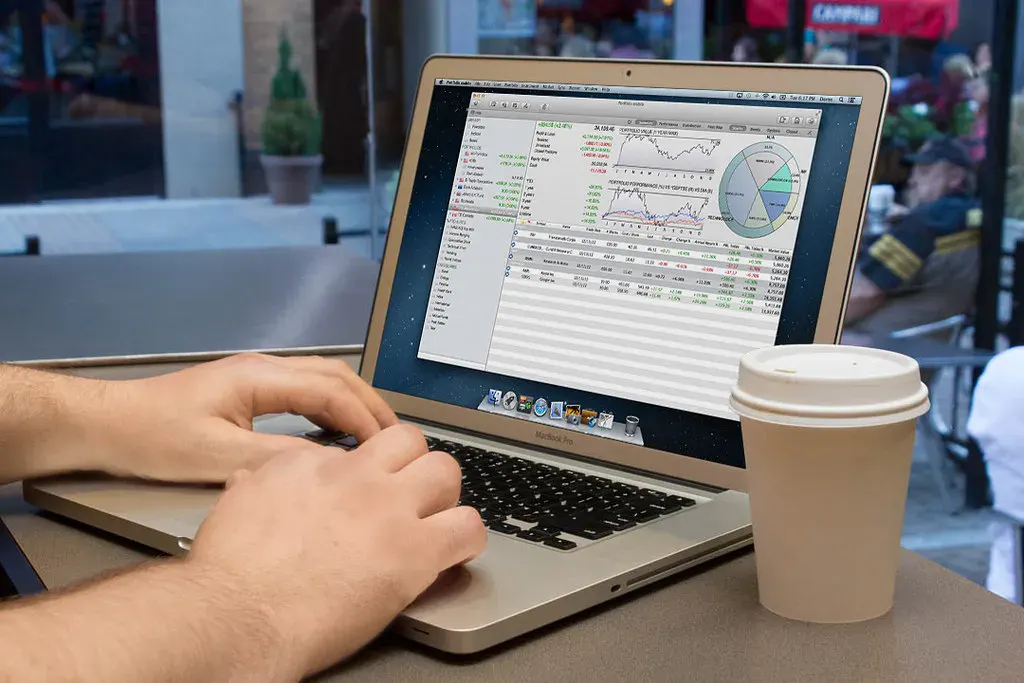 Close-up of a person's hands typing on a silver MacBook Pro laptop, which displays a financial application with various charts (line graphs, pie chart) and tables showing stock market data, including portfolio values and performance metrics. A white disposable coffee cup sits next to the laptop on a table, with a blurred outdoor cafe in the background.