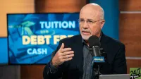 A bald man with a white beard and glasses, Dave Ramsey, is speaking into a black microphone with 'THE RAMSEY SHOW' branding. He is wearing a dark suit jacket over a blue-grey collared shirt and is gesturing with his right hand. Behind him, a large blue screen features blurred images of a graduation cap and money, with prominent yellow text displaying 'TUITION,' 'DEBT FREE,' and 'CASH.'
