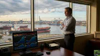 A man in a striped shirt stands with crossed arms, looking out a large office window at a busy port with cargo ships and cranes under an overcast sky. In the foreground, a desk holds a computer monitor displaying multiple financial charts with blue and white lines, a keyboard, mouse, and desk organizer.
