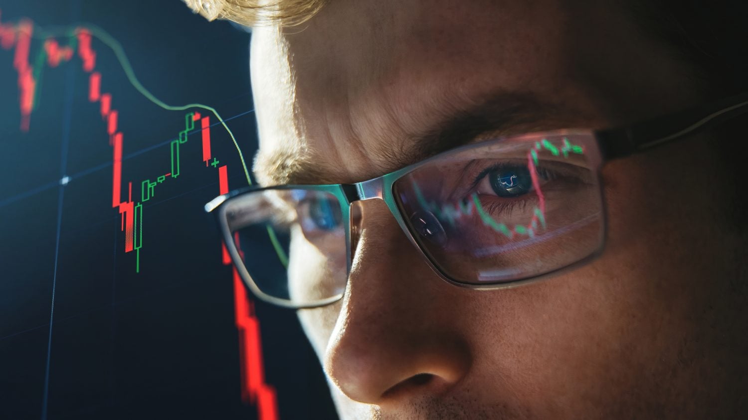 Close-up portrait of focused crypto trader analyst wearing eyeglasses looking at computer screen analyzing stock market charts. Eyeglasses reflection cryptocurrency downtrend charts. Bitcoin crash
