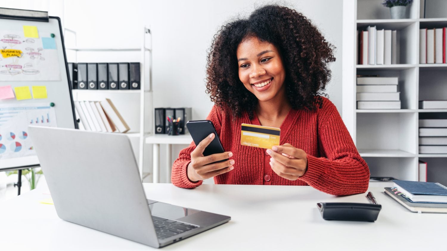 A woman is smiling while using her cell phone to pay for something. She is holding a credit card in her hand
