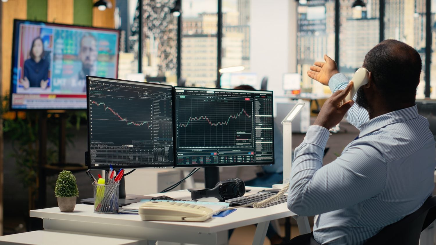 A Black man in a light blue striped shirt sits at a white desk in an office, seen from behind. He is holding a beige landline phone to his right ear and gesturing with his left hand. In front of him are two computer monitors displaying dark interfaces with colorful stock market charts and data. To his left, a smaller potted plant and a pen holder are on the desk. In the background, a large TV shows two news anchors, and outside large windows, a city skyline is visible.