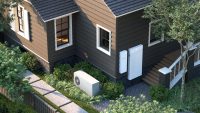 An overhead shot of a modern house with dark brown and dark grey siding, white window frames, and a dark grey shingled roof. In front of the house, a white Xera heat pump with a circular fan is visible among green shrubs and ornamental grasses. Mounted on the side of the house are two white rectangular units: a smaller one labeled "SPAN" and a larger, unmarked energy storage unit. A light-colored wooden staircase with white railings leads to an entrance on the right, and concrete stepping stones form a path through the green landscaping.