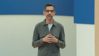 Sundar Pichai stands before a blue and white backdrop, wearing a grey button-up shirt and glasses, hands clasped together while speaking at Google I/O 2024.