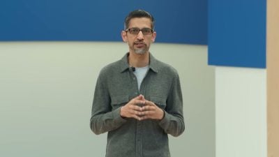 Sundar Pichai stands before a blue and white backdrop, wearing a grey button-up shirt and glasses, hands clasped together while speaking at Google I/O 2024.