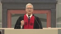 Tim Cook stands at a podium wearing black academic robes and a red ceremonial stole with university seals, raising a fist while speaking at MIT's 2017 commencement ceremony.