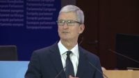 Tim Cook stands at a podium with microphones in front of a European Parliament backdrop, wearing a navy suit, white shirt, and dark tie while speaking.