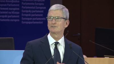 Tim Cook stands at a podium with microphones in front of a European Parliament backdrop, wearing a navy suit, white shirt, and dark tie while speaking.