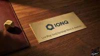 A rectangular brass plaque with the blue IonQ logo and the words "Live Blog: IonQ Earnings News & Analysis" engraved on it rests on a dark, polished wooden desk. To the left of the plaque, a faceted, irregular brass object sits on a dark red leather portfolio or notepad. The scene is lit by warm, directional light, creating distinct shadows and highlighting the metallic and wooden textures.