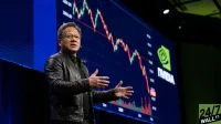 A man identified as Nvidia CEO Jensen Huang, wearing glasses and a black leather jacket, stands on a stage, gesturing with his hands as if speaking. Behind him, a large blue screen displays a stock chart with dramatic red and green lines indicating price fluctuations, along with the bright green NVIDIA logo and 'NVIDIA' text. Numbers '3200.00' and '3000.00' are visible on the chart's vertical axis. The bottom right corner features a '24/7 WALL ST.' watermark.