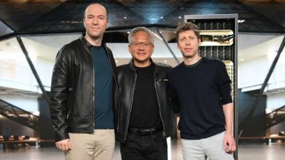Three men stand side-by-side, smiling towards the camera. In the center, NVIDIA CEO Jensen Huang, an East Asian man with gray hair and glasses, wears a black leather jacket over a black shirt and dark pants. To his left, a bald Caucasian man wears a black leather jacket over a dark teal long-sleeve shirt and light beige pants. To Huang's right, a Caucasian man with curly brown hair wears a dark navy long-sleeve shirt and light gray pants. The background features modern architectural elements, including glass panels and dark structural beams, with a server rack visible on the right.