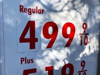 A close-up shot of a white gas station price sign with large red numbers. The word 'Regular' is at the top, followed by the price '499 9/10' in bold red. Below it, the word 'Plus' is visible, with partially seen red numbers below. Shadows from tree leaves fall across the bright white sign, partially obscuring the numbers.