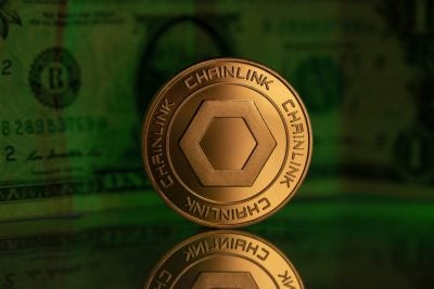 A golden Chainlink cryptocurrency coin is prominently displayed, standing upright on a reflective surface. The coin features a central hexagonal design and the word 'CHAINLINK' embossed around its circular edge. The background is a soft, blurred green, subtly revealing elements of a US dollar bill.