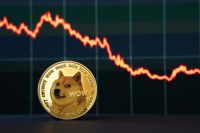 A close-up shot of a physical gold-colored Dogecoin cryptocurrency coin in the foreground. Behind it, a dark digital screen displays a red, fluctuating line graph that trends significantly downwards, indicating a market decline. The screen has a subtle grid pattern.